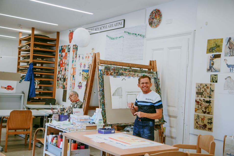 Ein Kunststudio mit zwei Personen. Eine Person sitzt an einem Tisch voller Kunstmaterialien und malt. Die andere Person steht neben einer Staffelei und hält ein Stück Papier, während sie lächelt. An den Wänden hängen bunte Kunstwerke und Zeichnungen. Künstler in einem Atelier, der an einer Staffelei arbeitet, während eine andere Person an einem Tisch sitzt und malt. Kunstwerke schmücken die Wände.